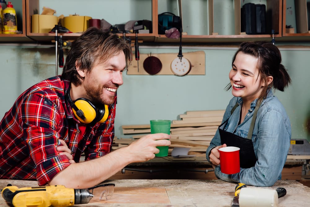 Man and girl carpenters having break from indoor work in wood workshop. Coffee time, drink, share experiences, discuss. Teacher and student. Laughing smile.